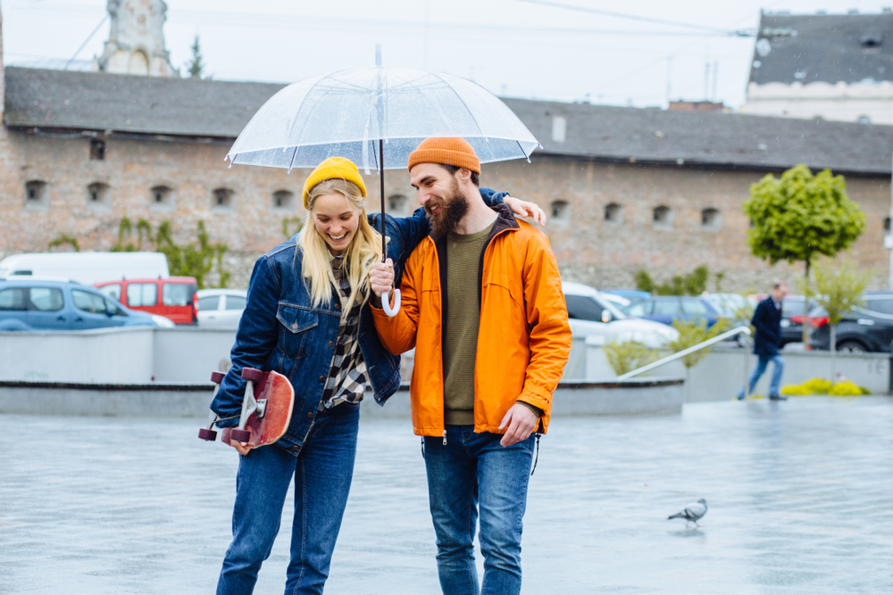 A couple in thicker jackets huddle under an umbrella not worrying about what to wear in London in February as they were prepared for the rain with their clear umbrella and yellow and orange beanies. 