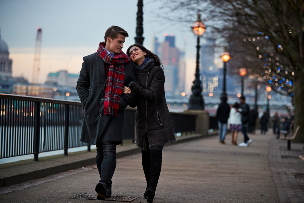 A couple walks down the Thames River, not worrying about what to wear in London in February as they are bundled with scarves and jackets to keep warm.