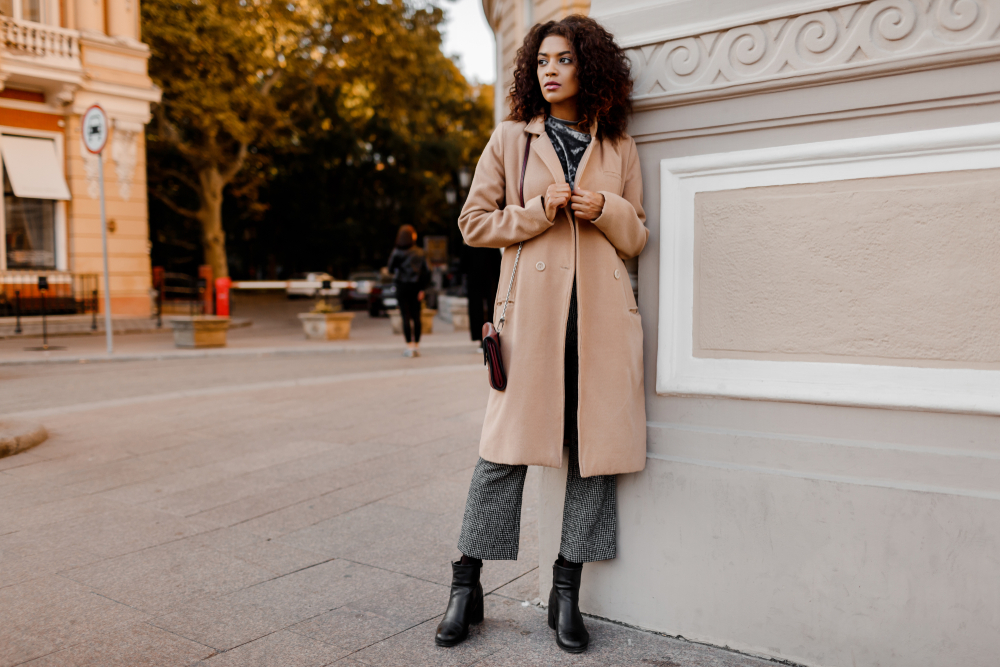 A women leans against a monument in ankle boots, trousers, and a long, tan wool coat. 