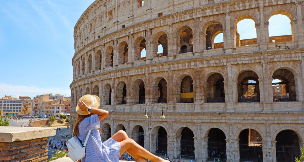 woman sitting in front of a building in Rome italy wearing a blue dress with her hand on her tan hat