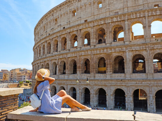 woman sitting in front of a building in Rome italy wearing a blue dress with her hand on her tan hat