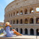 woman sitting in front of a building in Rome italy wearing a blue dress with her hand on her tan hat