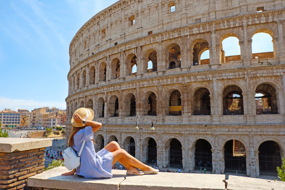 Woman sitting on a brick wall wearing a sun hat, small backpack, and short, blue dress, showcasing what to wear in Rome.