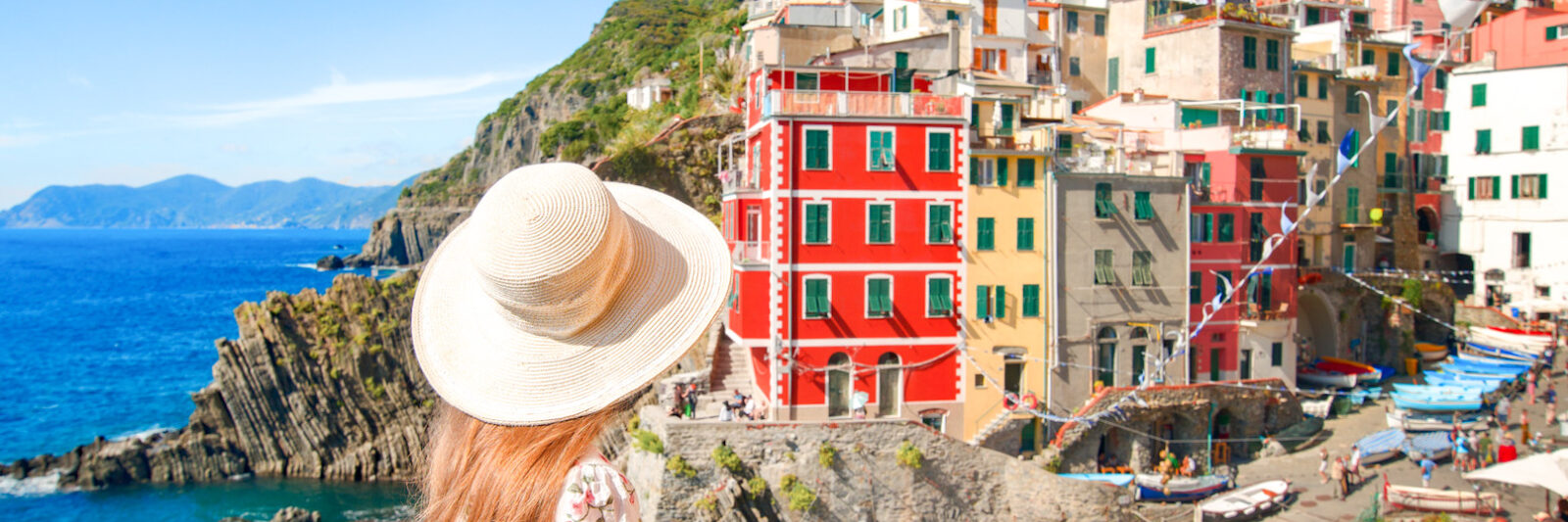 woman sitting on the edge of a brick wall wearing a white floral dress and sun hat overlooking lots of colorful buildings cascading down the side of a small hill with some water in behind the woman