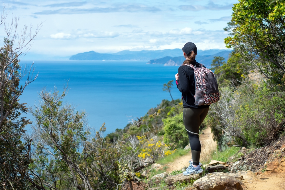 Woman on a hiking trail overlooking the ocean wearing leggings, a light jacket, ball cap, backpack, and hiking shoes.