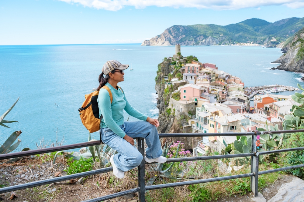 Woman in jean and a long-sleeved shirt sits on a metal railing overlooking Cinque Terre.