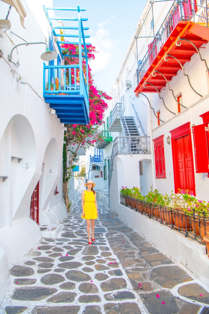 Woman in a short, yellow dress walking down a colorful alley in Greece with lots of flowers.