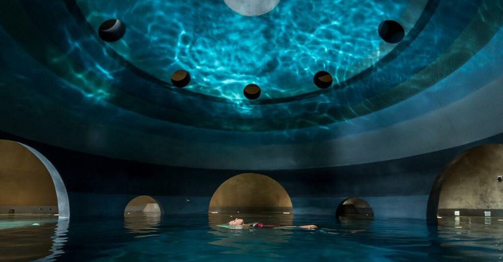 person floating on their back in a spa with giant gold balls around them and reflection of water on the celling in a round room