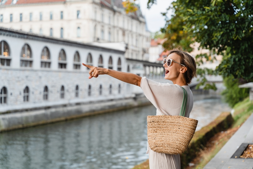 A woman points across a river in Paris: she wears sunglasses, a slowly dress, and has a woven bag on her shoulder. 