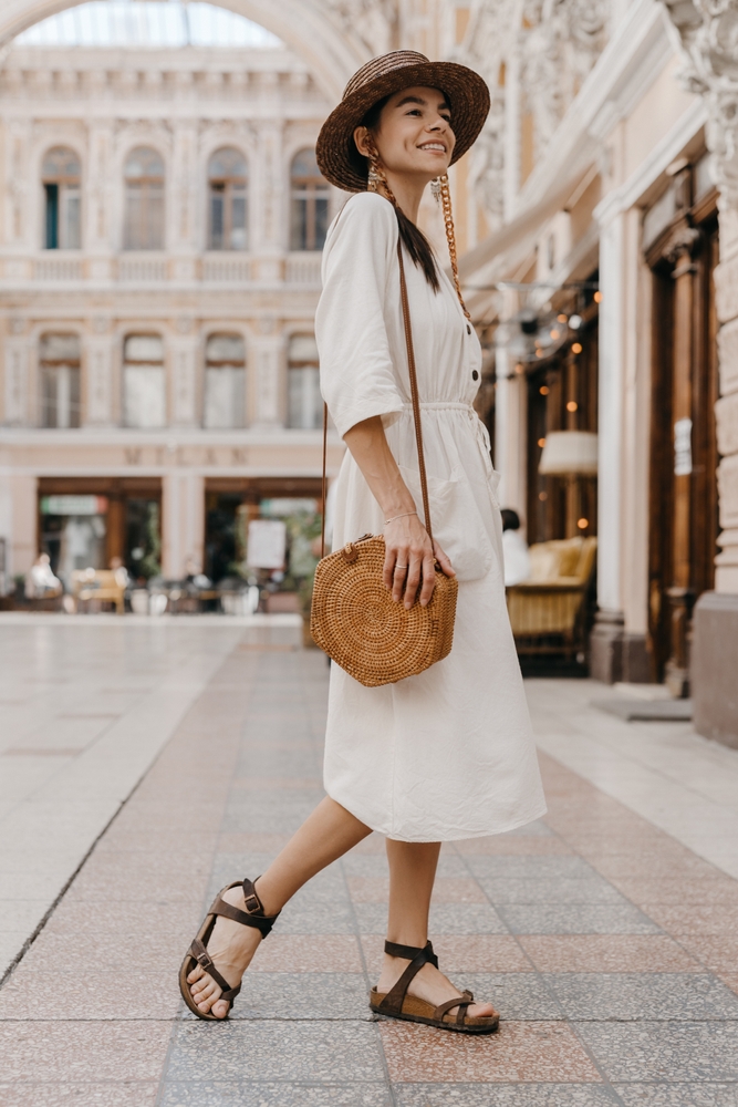 A woman in a linen white dress and wove crossbody bags stands and smiles in the middle of a European city. 