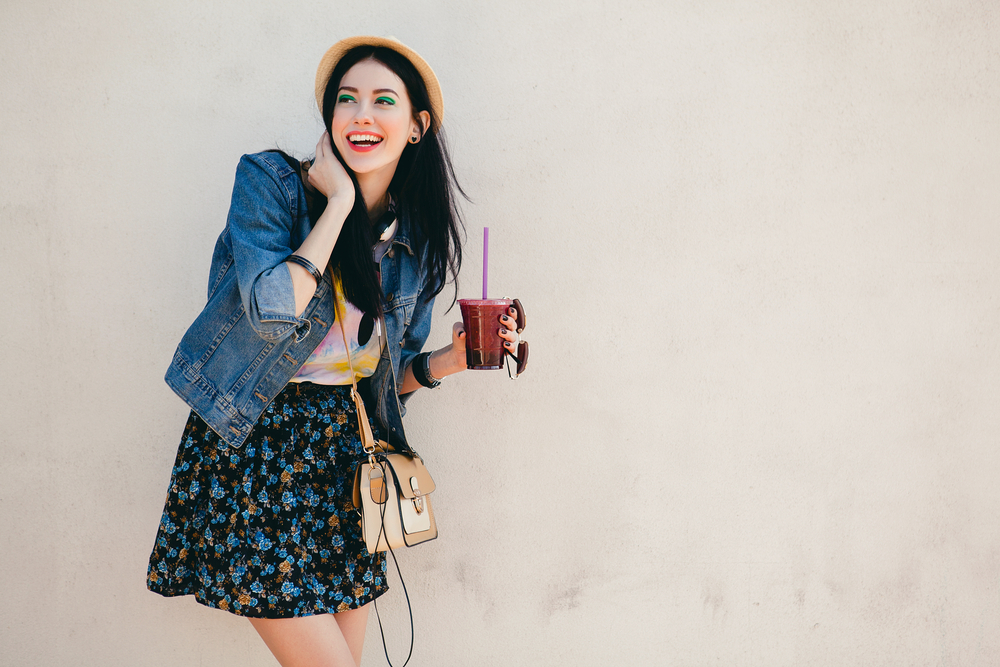 A girl smiles while enjoying a smoothie, comfortable in what she chose to wear when wondering what to wear in Europe in Summer: she dawns a jean jacket, a crossbody bag, a graphic t and a skirt. 