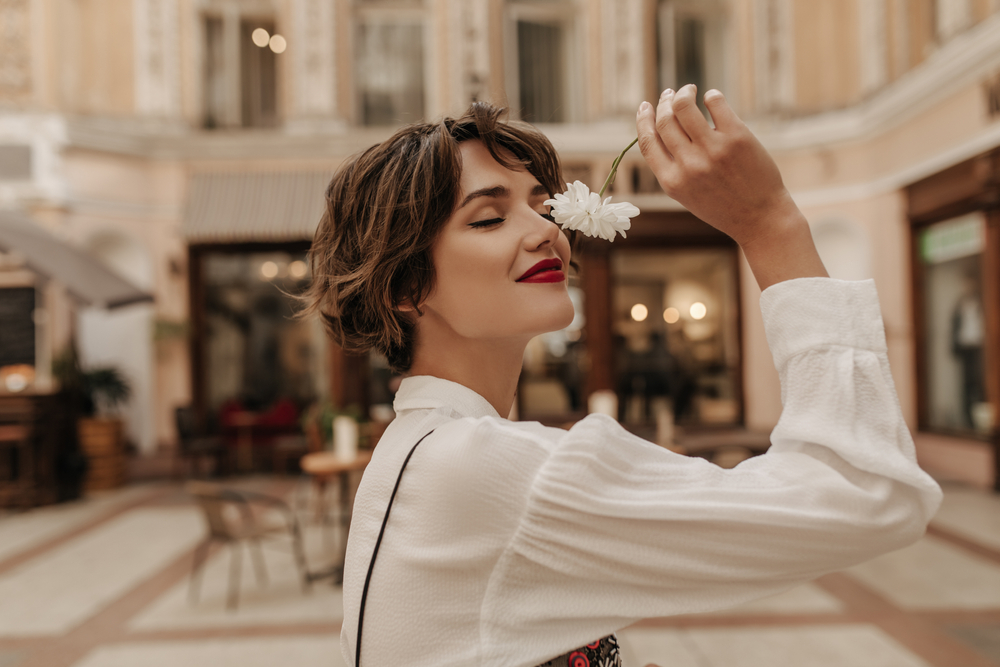 In a museum, a woman wears a white button down sheer shirt and she brings a white flower to her nose to smell it with a smile. 