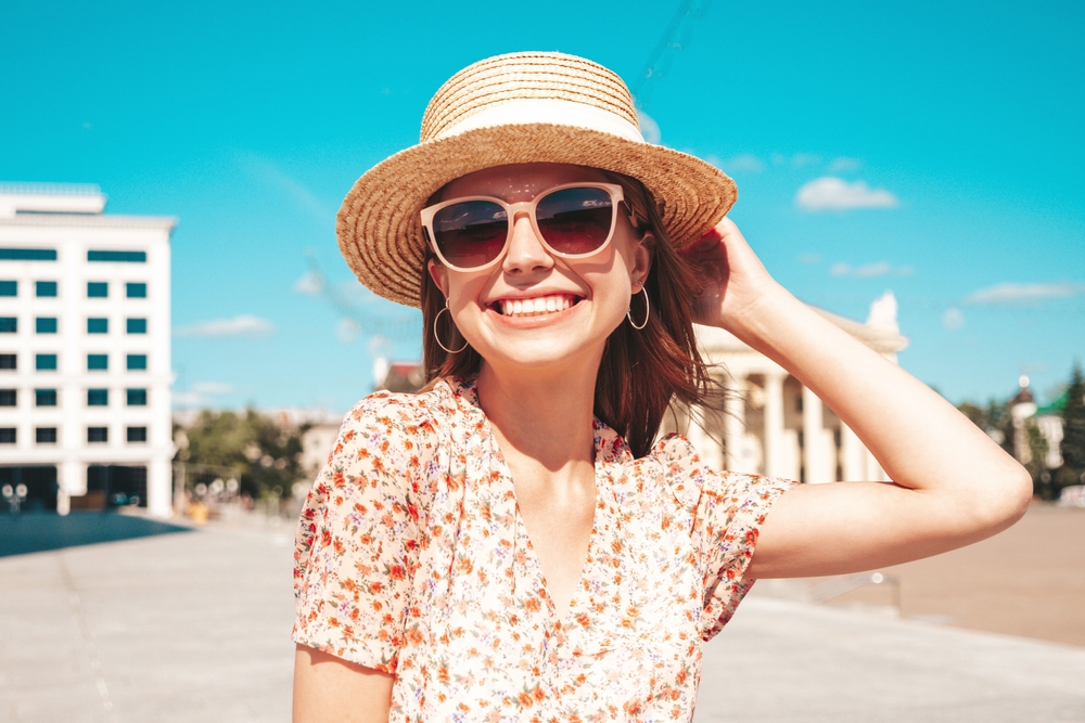 A woman in a straw sun hat and sunglasses smiles at the camera: she wears a floral summer dress. 