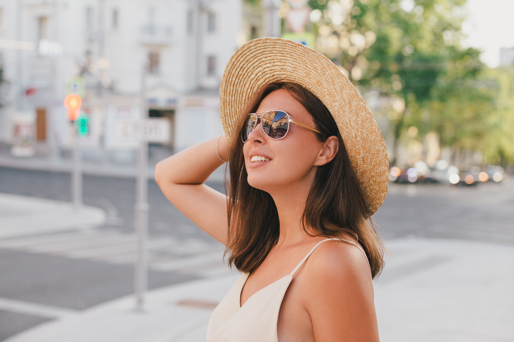 A woman smiles and looks up while wearing a comfortable tank top, a sun hat, and sunglasses, all perfect for what to wear in Europe in Summer. 