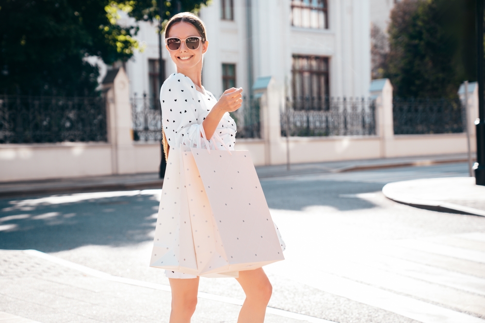 This woman wears sunglasses and a low bun as she shops through European streets: she wears a midi dress that is white with black hearts on it. 