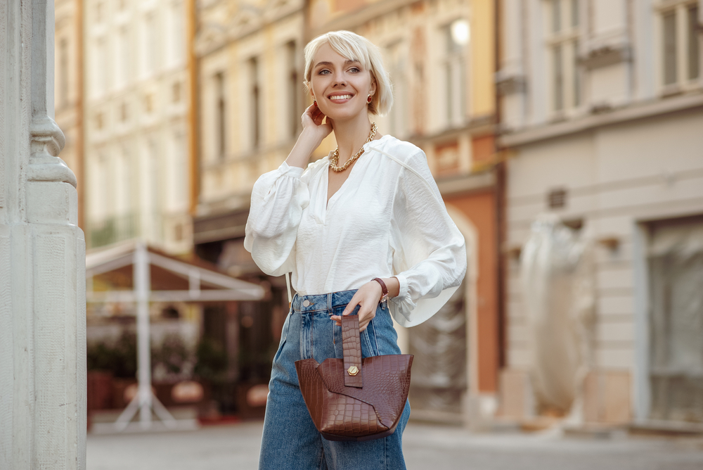 A woman with blonde short hair enjoys what to wear in Europe in Summer and keeping it casual with a button down white shirt and blue jeans, as well as a brown leather bag. 