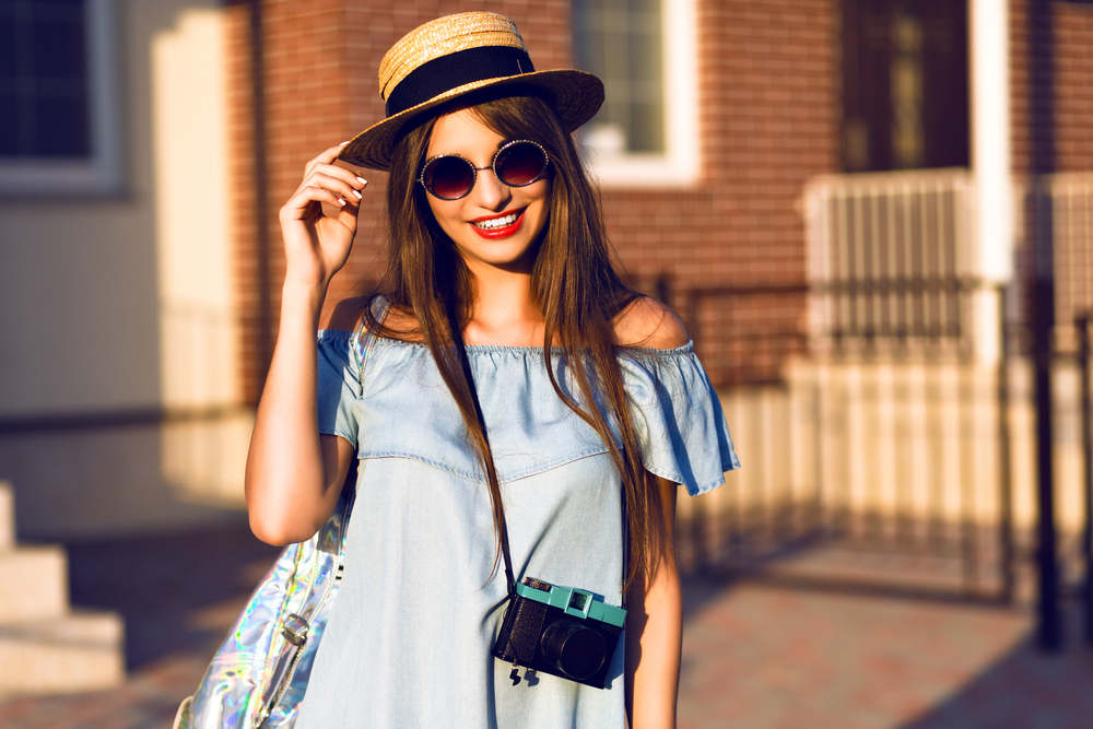 A woman smiles at the camera wearing an off-the-shoulder top and sunhat and glasses, which is perfect for what to wear in Europe in Summer. 