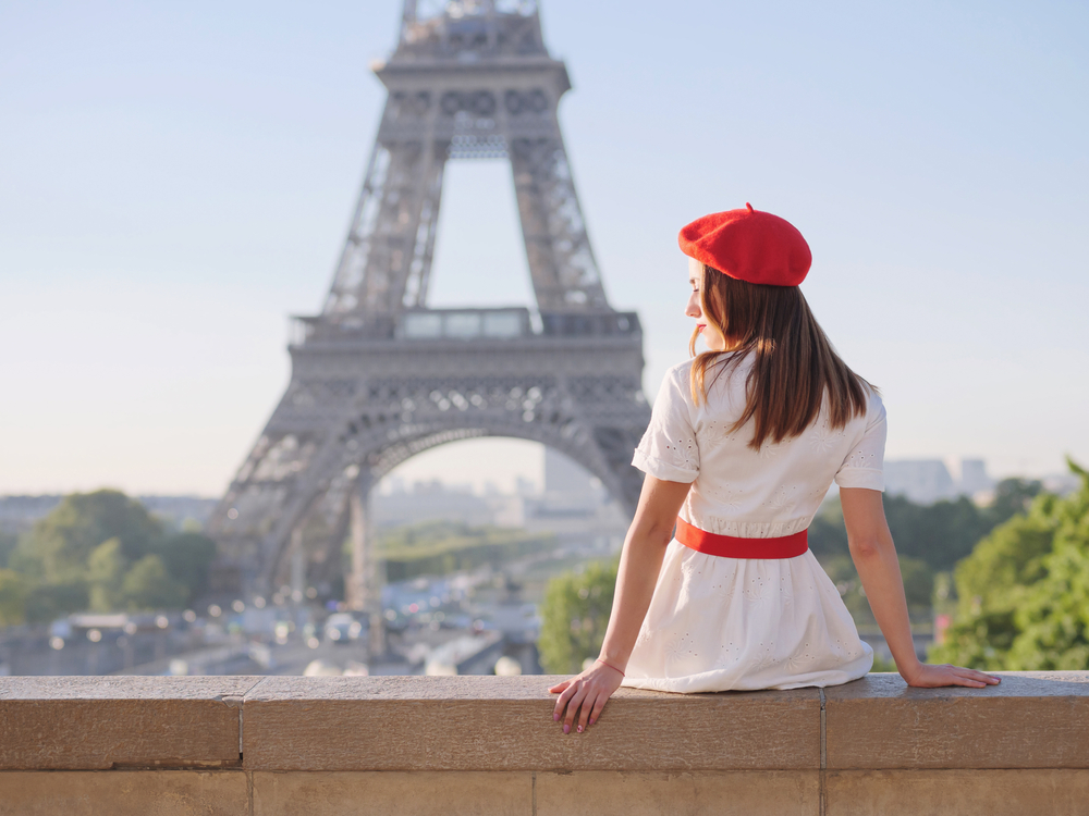 A woman sits on the edge, overlooking the Eiffel Tower: she wears a red beret, a red belt and a white sundress. 