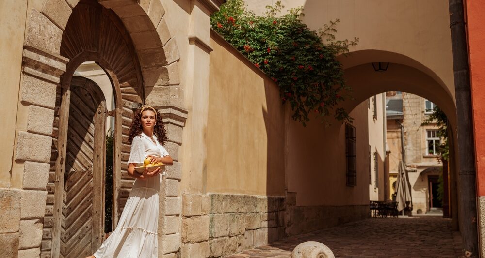 woman standing in a door wearing a long white dress, brown purse, and white had