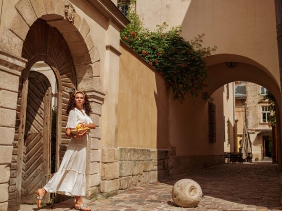 woman standing in a door wearing a long white dress, brown purse, and white had