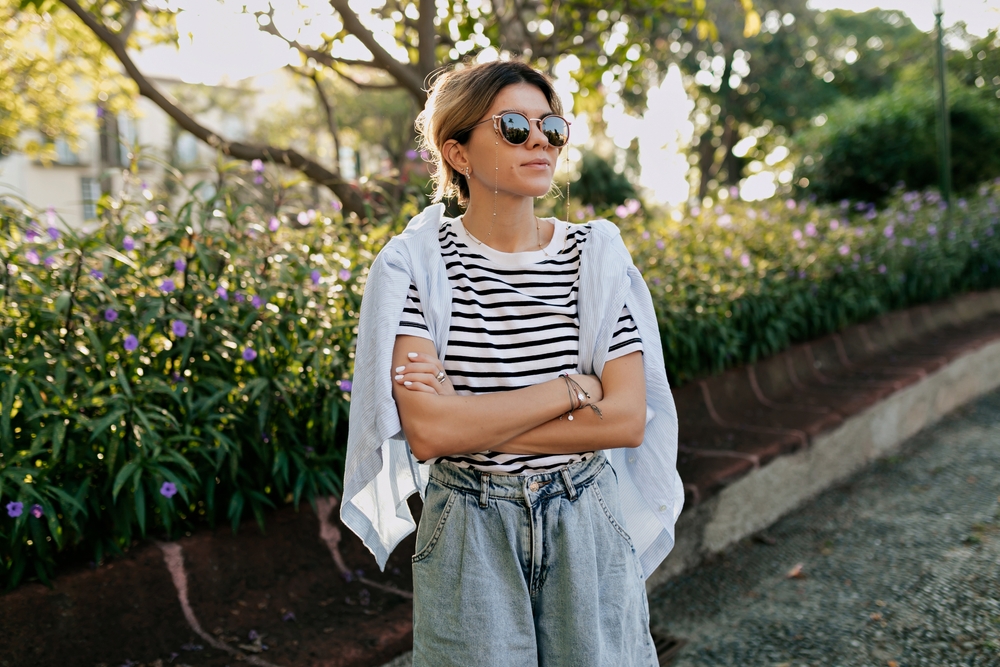 A woman stands in a park with a striped shirt on, statement sunglasses, a cardigan over her shoulders, and jeans. 