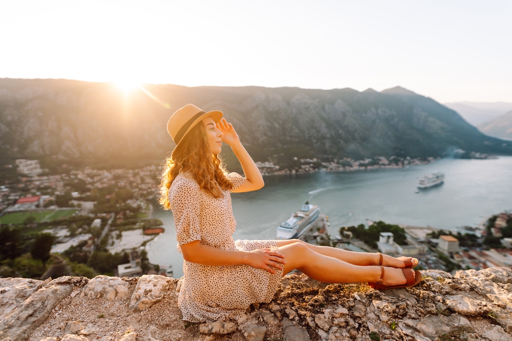 A woman sits on the coast of the water, on the rocks, not worrying about what to wear in Europe in Summer, as she is comfortable in her sandals, summer dress and hat. 