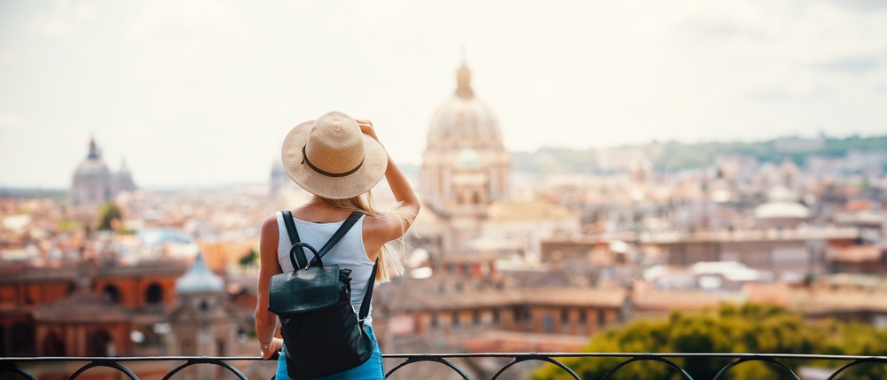 A women overlooks the views in Rome in a white tank top, blue shorts, a sun hat, and a leather backpack. 