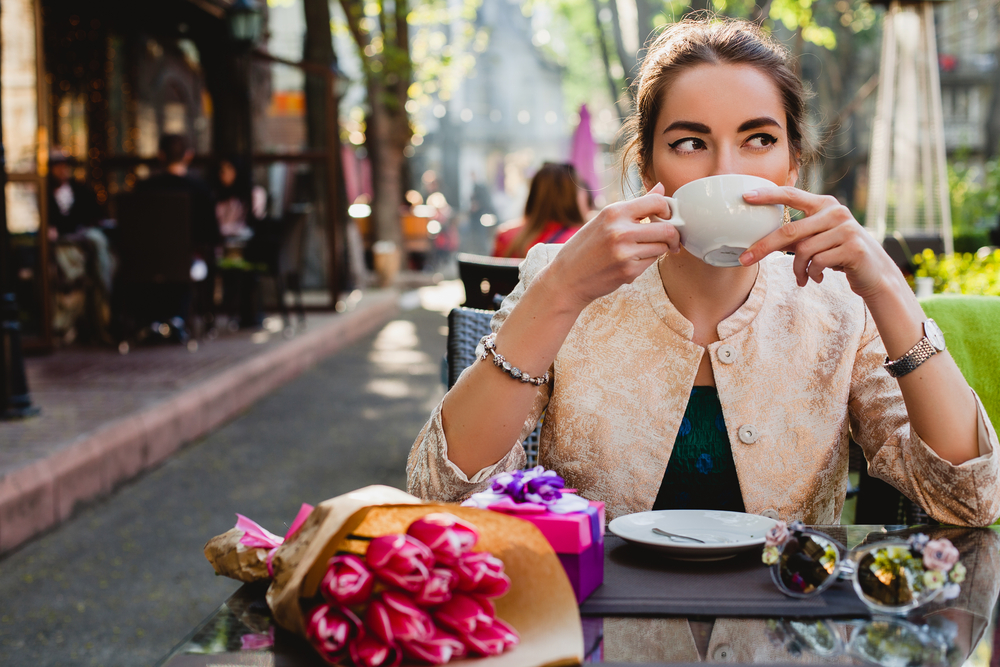 A woman sits at a table in Europe, sipping on coffee or tea, wearing a black t-shirt and a light cardigan as the sun goes down. 