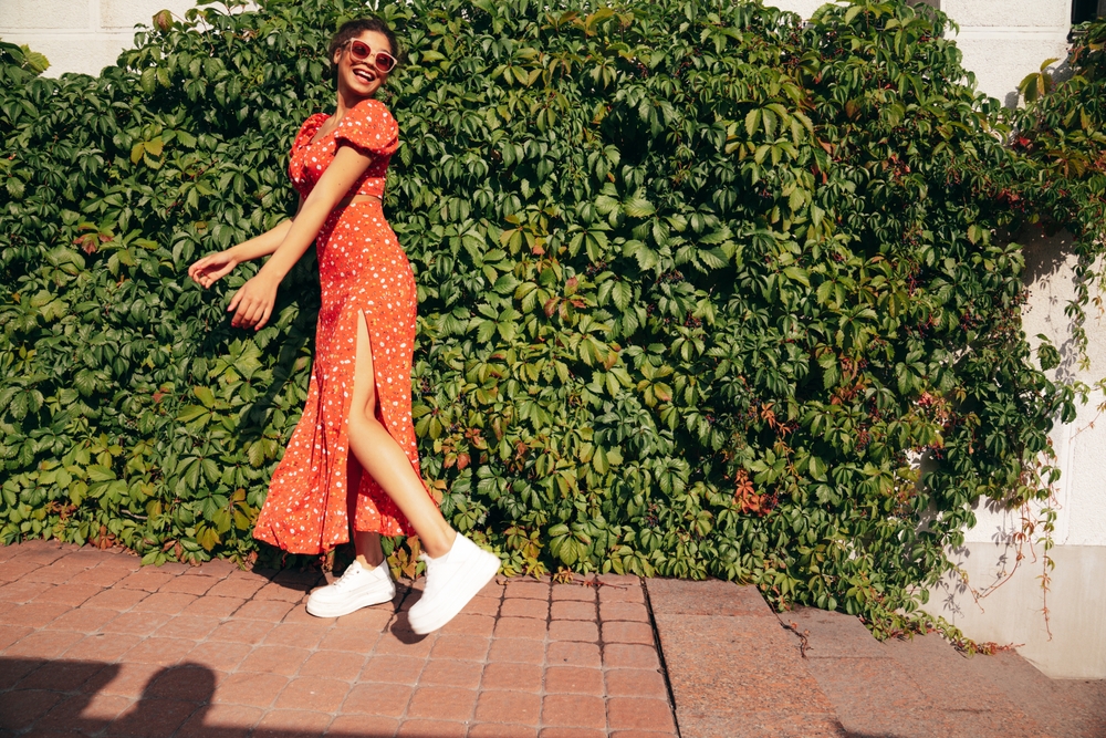 A woman poses against a wall that is covered in plants! She wars a sundress with a slit and comfortable sneakers. 