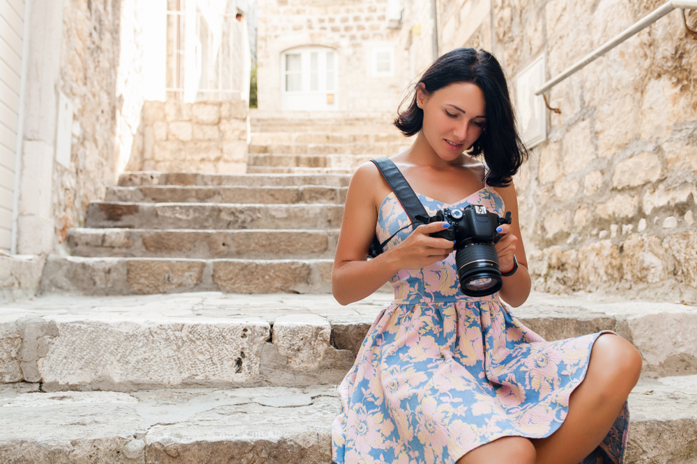 A woman sits on the edge of cobblestone stairs, looking at her camera: she wears a floral summer dress, cotton, which is perfect when considering  what to wear in the city in the summer. 