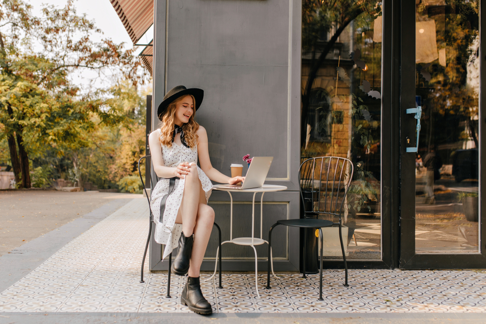 A woman perches outside a cafe in boots, a polka dot dress and a hat, working on her computer, not worrying about  what to wear in the city in the summer. 