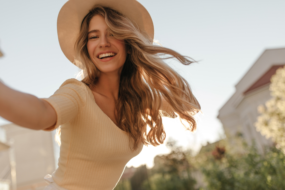 A woman smiles at the camera and her hair flows in the wind, matching her beige ribbed shirt and sun hat: in the background you can see elements of trees and the city. 