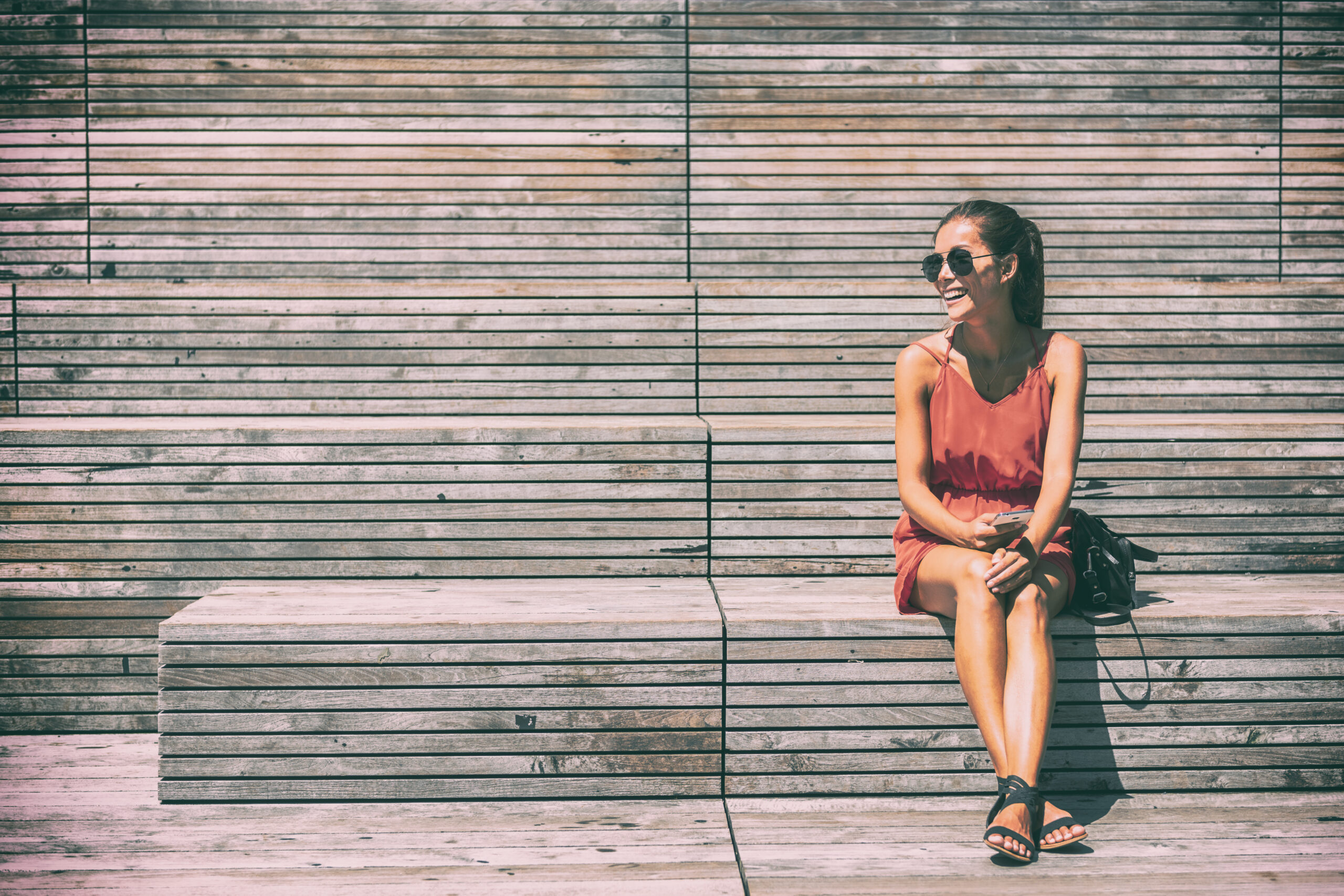 A woman sits and takes a break on a wooden bench, not worrying about  what to wear in the city in the summer because she is comfortable in her red/pink romper, with her sandals and crossbody bag. 