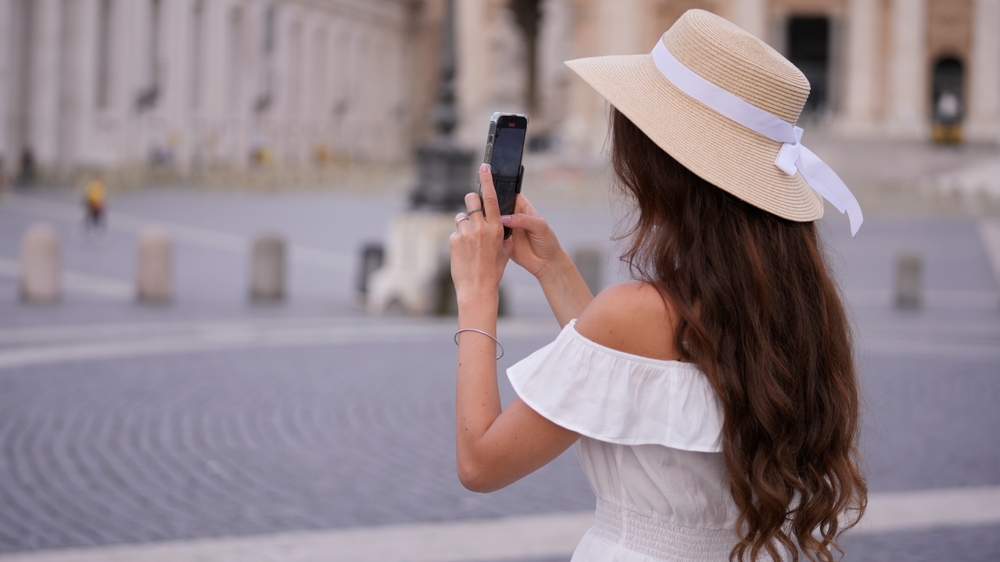 A woman in an off-the-shoulder-linen blouse matches her white outfit with her hat, as her straw hat has a white bow on it! She takes a photo on her phone of the city beyond the lens. 