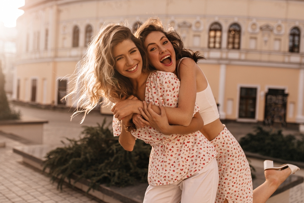 Two friends don't worry about  what to wear in the city in the summer: they are matching! As they hug, one wears contton pants and a floral blouse, while the other wears the floral pattern skirt and a white tank top. 