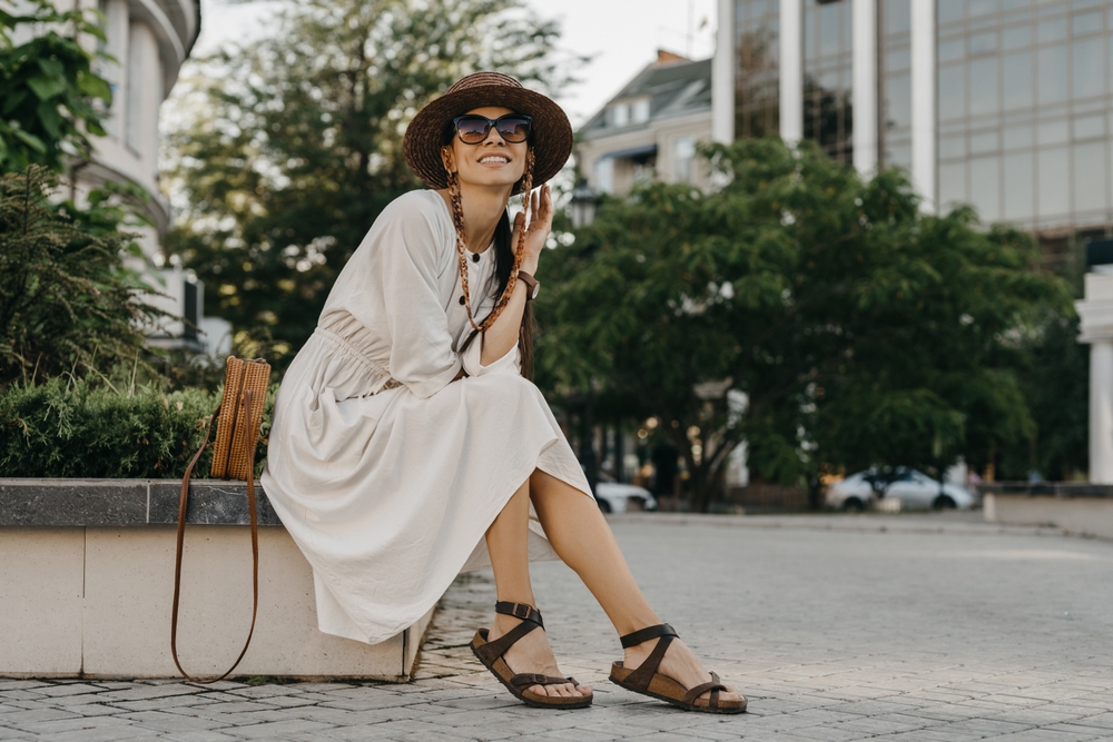 What to wear in the city in the summer is never an issue if you pack classics like this model: she dawns a beige maxi dress with comfortable sleeves and sandals. Her crossbody woven bag adds a chic element as she sits on a ledge. 