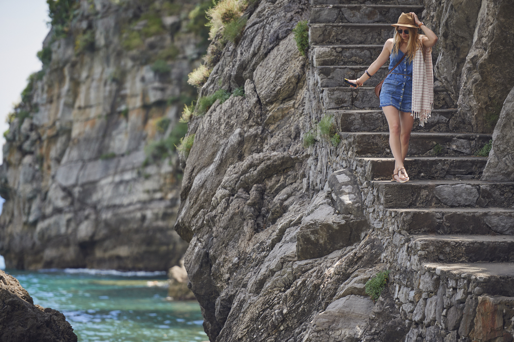 a women in jean dress, sunhat, lightweight scarf, crossbody bag and sandals walking down stairs to beach