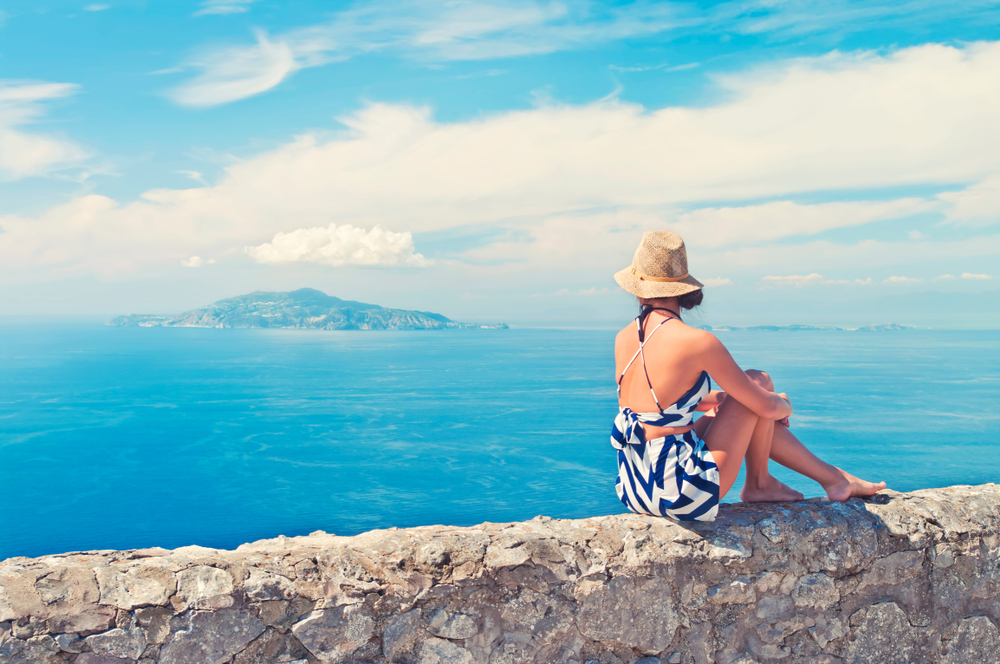 a woman in a matching blue and white linen shorts set sitting on a wall overlooking the ocean 