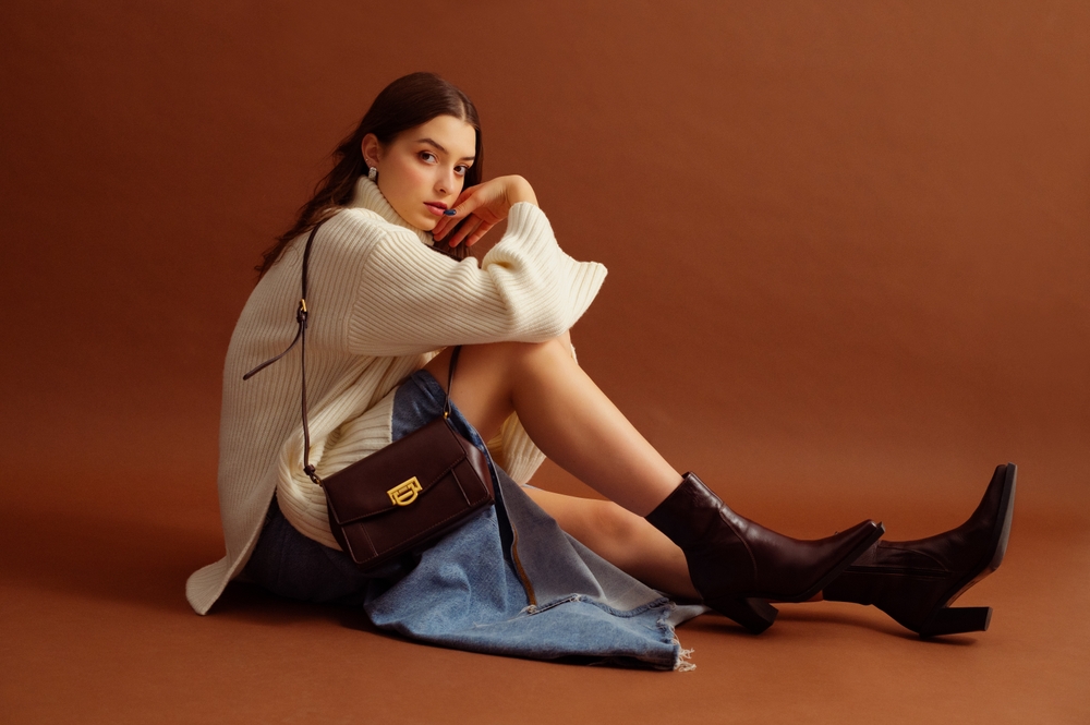 a woman sitting on ground in a creme turtleneck, jean skirt and heeled boots