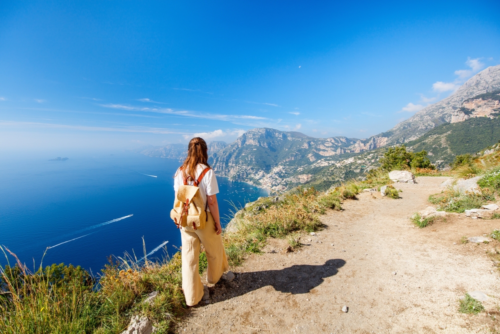 a woman in linen pants and tee shirt with a backpack overlooking an arial view of Amalfi Coast