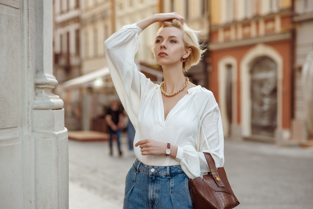 A woman runs a hand through her hair as she wears a white blouse and high waisted jeans with statement jewelry. 