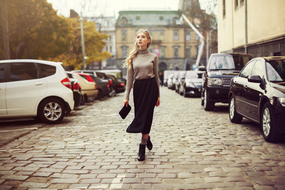 A woman walks down cobblestone streets in a black skirt and ankle boots and turtle neck shirts. 