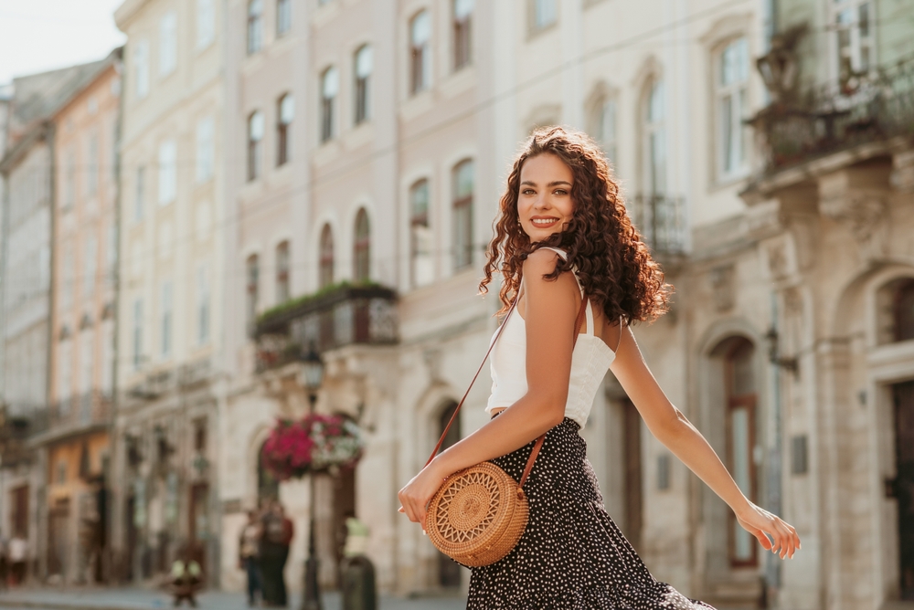 A woman in a white tank top and polka dot skirt twirls in the street of Europe with a straw crossbody bag. 