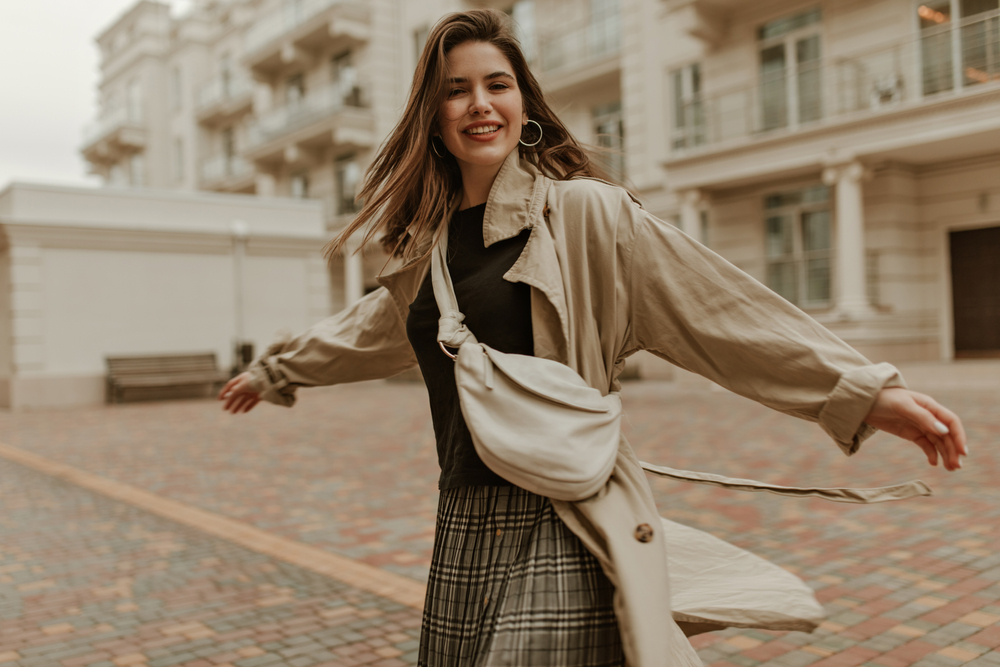 A woman doesn't wonder what to wear in Europe in Spring, as she is ready with a trench coat, a plaid skirt, a black top and a crossbody bag. 