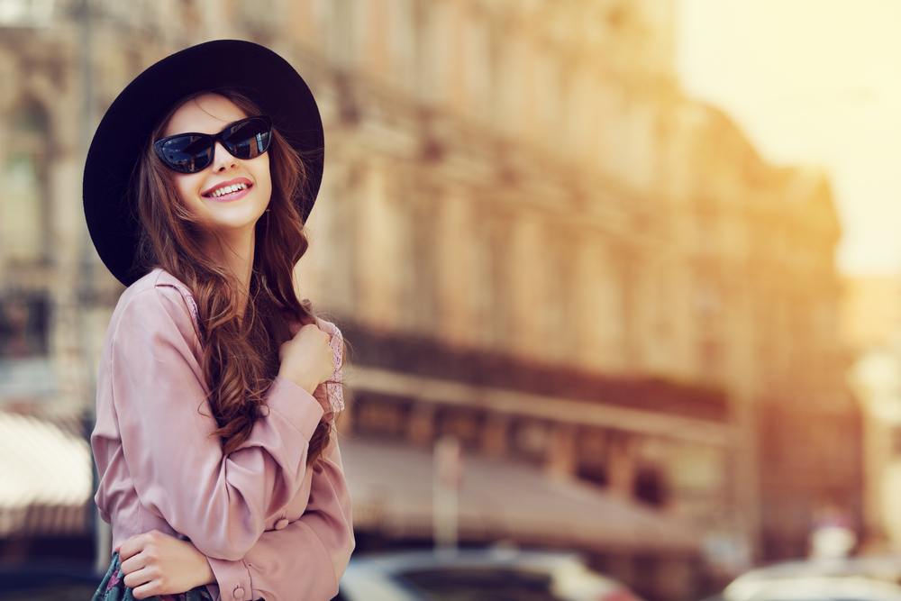 A woman smiles at the camera while enjoying Europe: she wears a pink blouse, sunglasses and a large sunhat. 