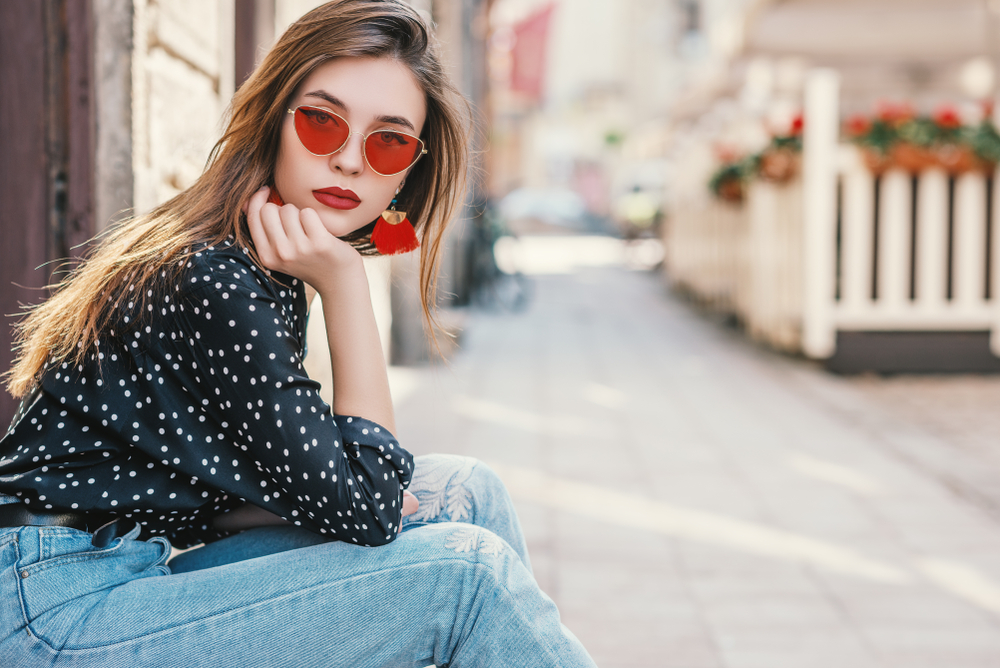 A woman in red sunglasses, a polka dot blouse and jeans sits on a curb in Europe and looks at the camera. 