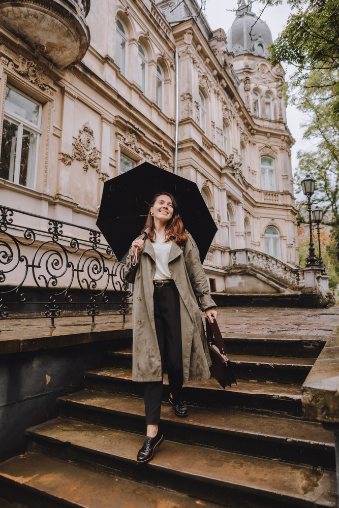 A woman in a trench coat, white shirt and black pants walks down some stairs, crying an umbrella as she walks though Europe. 