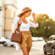 woman wearing a cute outfit for spring in europe standing on the sidewalk. she is wearing brown pants and a white top and holding a brown bag