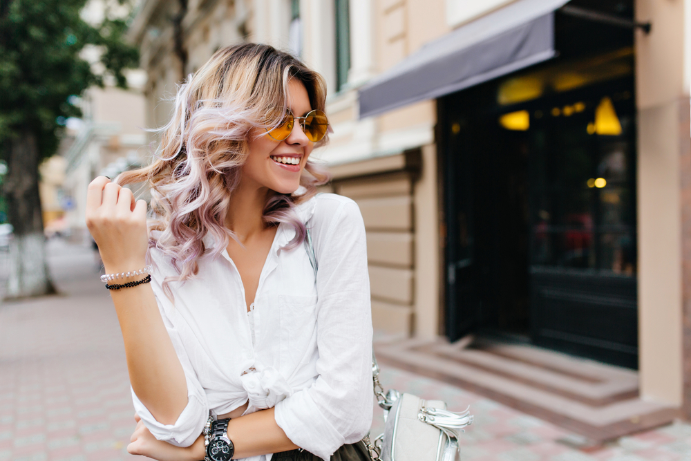 A woman twirls her hair in Europe in a white button downed yellow sunglasses in the street. 