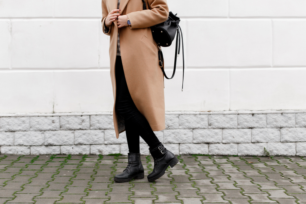 A woman stands by a building in a long wool coat, thin black jeans and boots, ready for the cooler weather in fall in New York. 